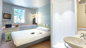 Een hotelkamer met een bed, een bureau met een laptop, een televisie, een raam met uitzicht op bomen, en een badkamer met een wastafel en spiegel.