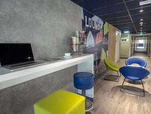 A lounge area with a laptop on a counter, a bookshelf, and colorful chairs. - ibis budget Brignoles Provence Verte (Brignoles)