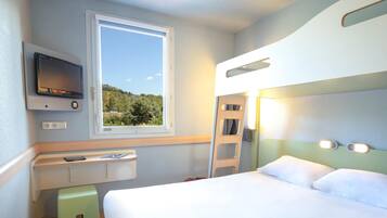 A compact hotel room with a bed, a small desk with a TV, a window with a view, and a ladder.