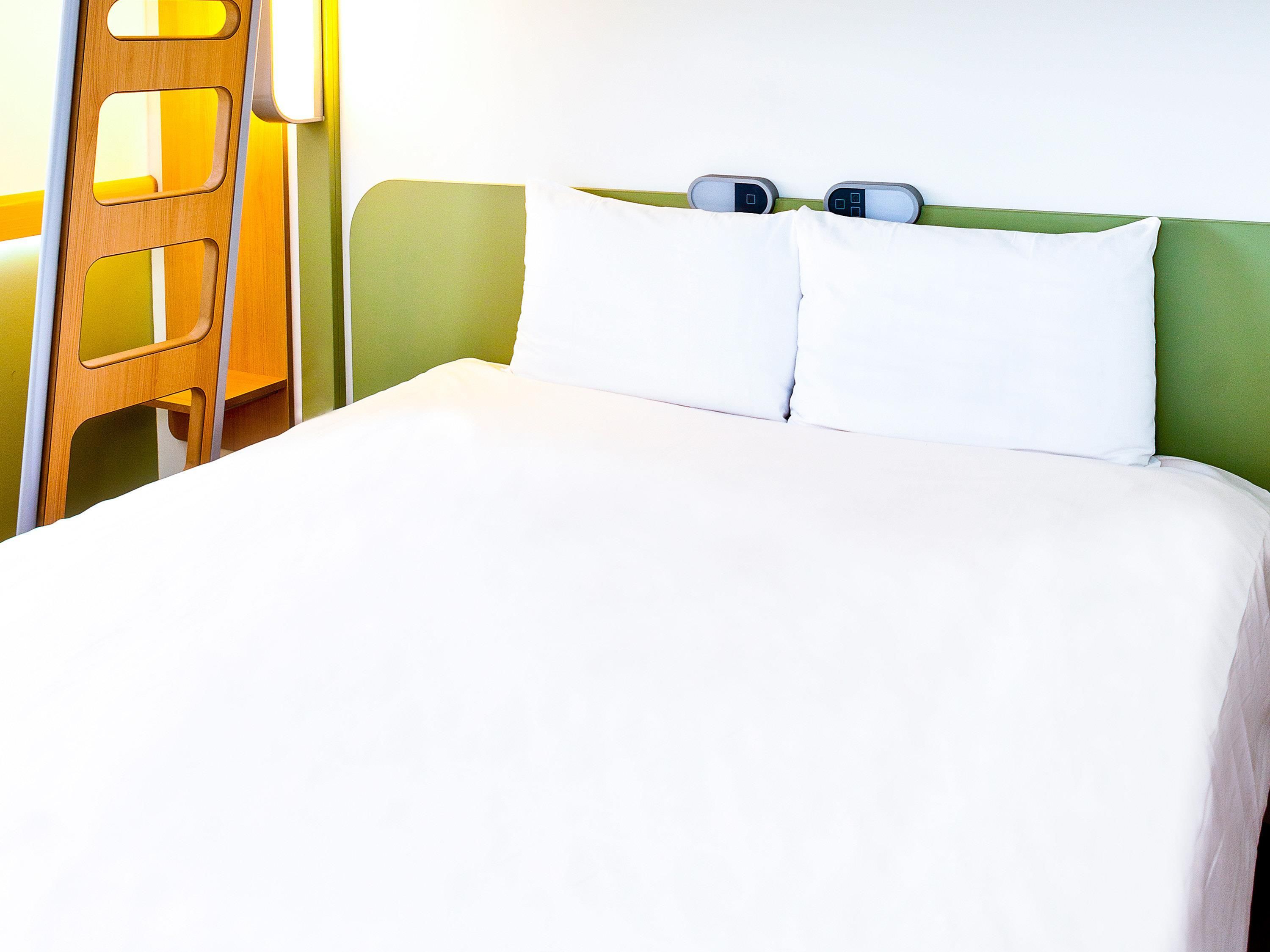 Cama com roupa de cama branca, cabeceira verde e dois relógios de cabeceira.