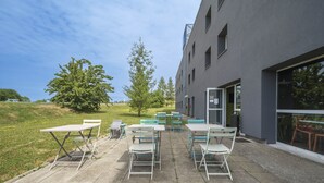 Un espacio al aire libre con mesas y sillas, un edificio con amplias ventanas y un campo verde al fondo.