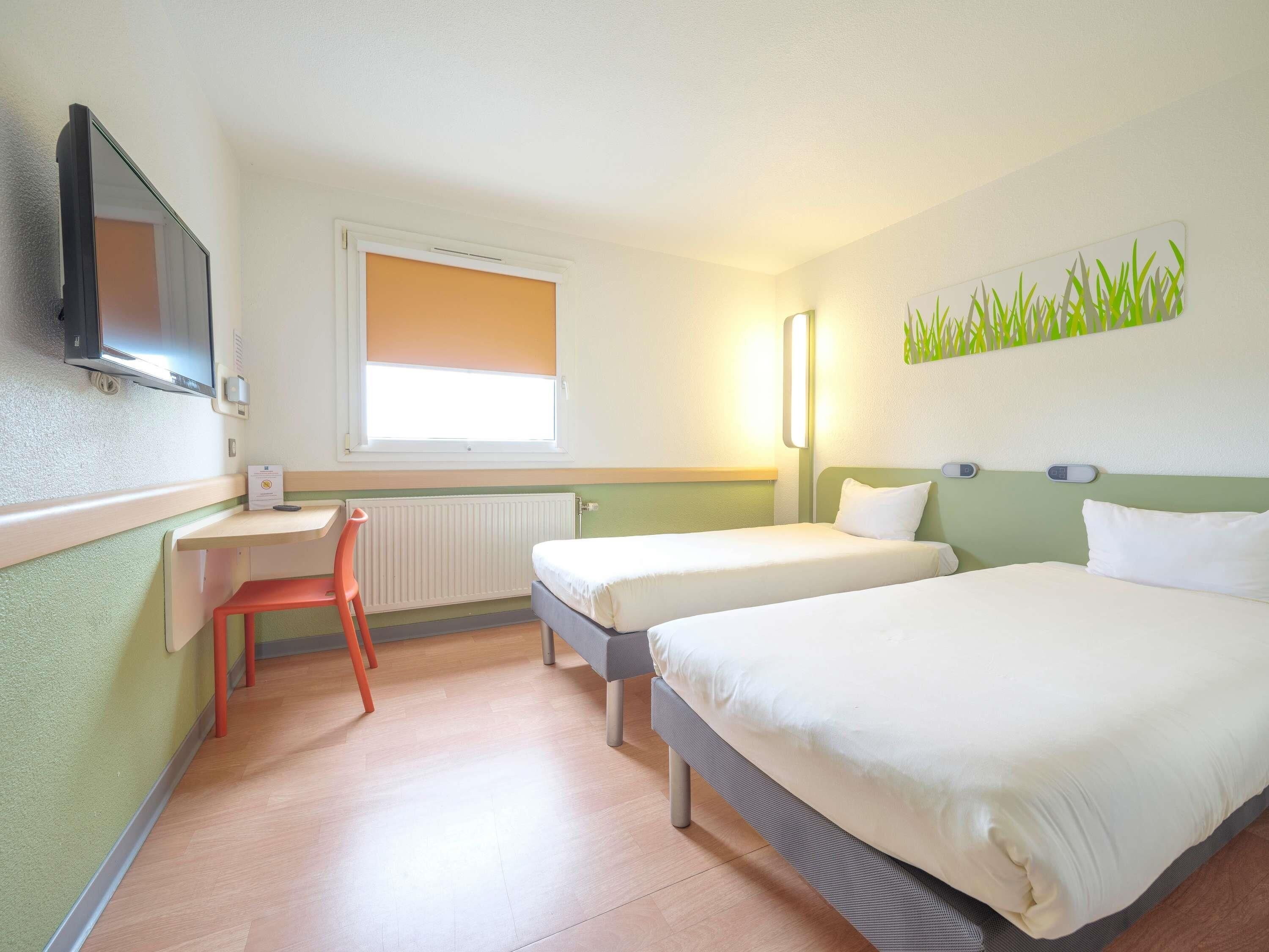 Une chambre d’hôtel avec deux lits, un bureau, une chaise, une télévision et une fenêtre avec des stores.