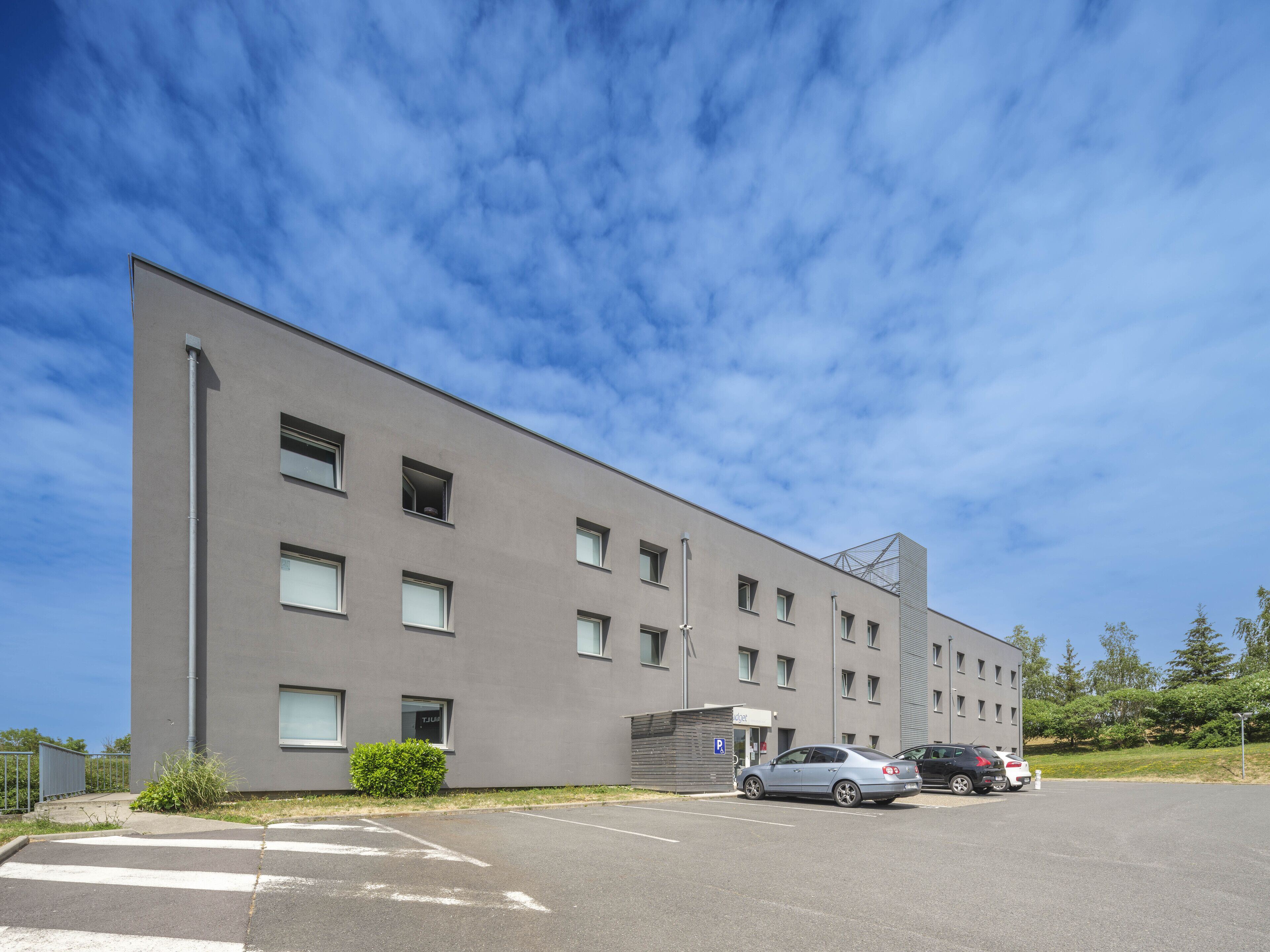 A modern building with a flat facade, multiple windows, and a parking area with cars.