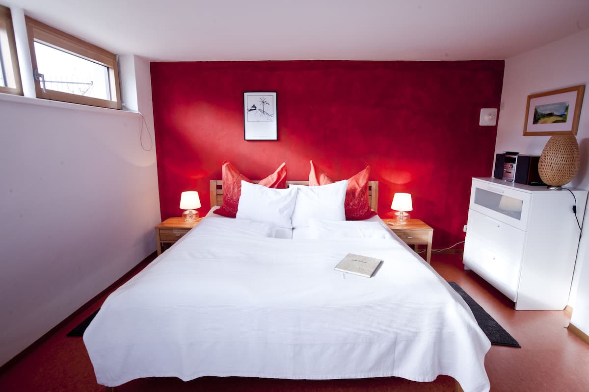 A neatly made bed with white linens and red pillows, a bedside table with a lamp, and a small cabinet with a lamp on top.