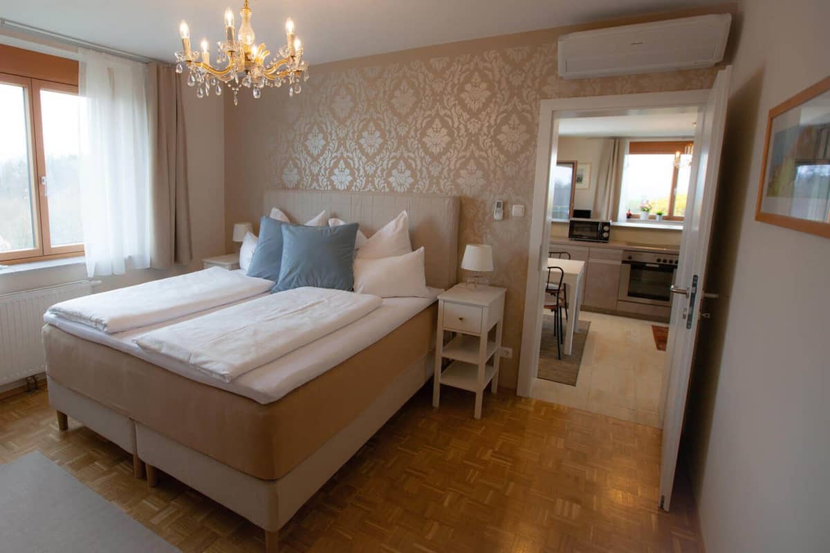 A bedroom with a double bed, a bedside table, a nightstand, a chandelier, a window with curtains, a radiator, and a view into the kitchen.