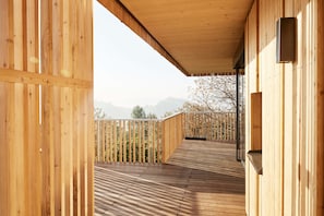 A wooden deck with a railing, a view of trees and mountains, and a building with a sloped roof.