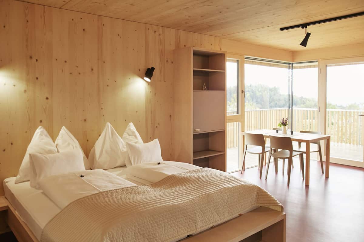 A bedroom with a wooden headboard, a bed with white bedding, a bedside table, and a view of a balcony with a table and chairs.