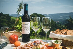 A bottle of wine, two glasses, an apple, an orange, a basket of bread, and a bowl of salad on a table with a scenic view in the background.
