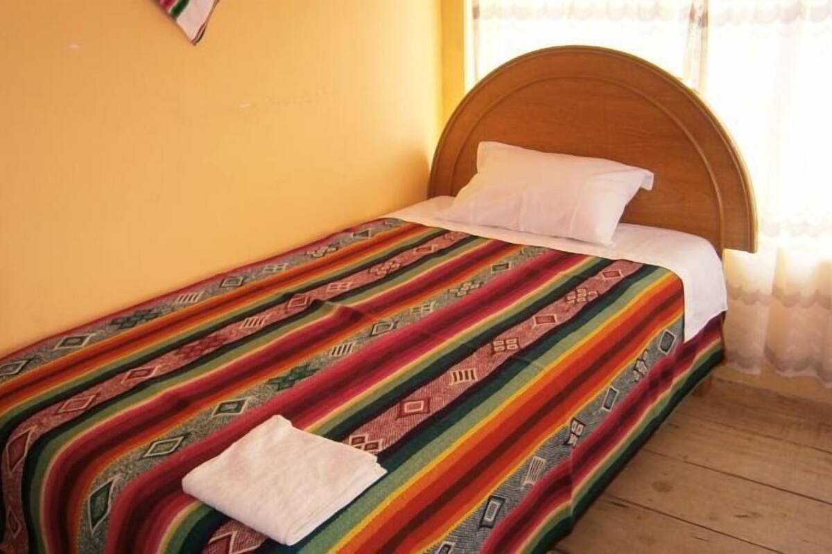 A single bed with a colorful blanket and a pillow in a room with wooden flooring and a yellow wall.