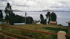 A view of a lake with mountains in the background, a wooden fence in the foreground, and a few buildings near the water. - Samay Wasi Salakancha (Isla Amantani)