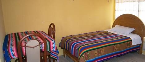 Una habitación pequeña con una cama individual y una silla, ambos cubiertos con mantas coloridas.