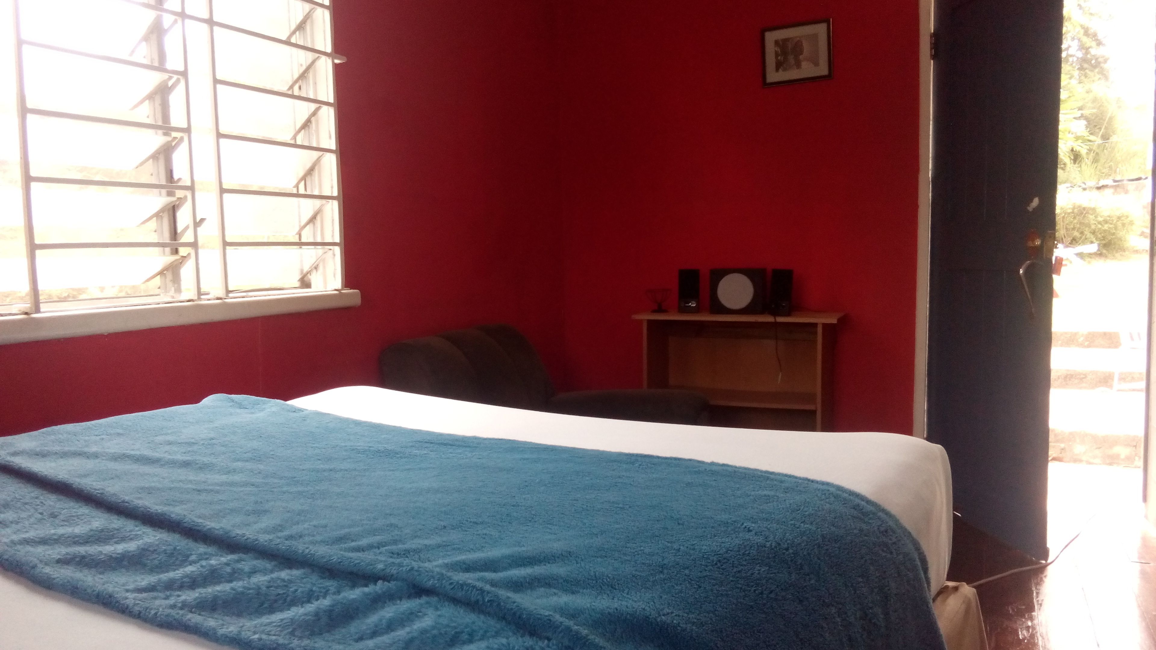 A room with a bed, a window, a red wall, a small table with a speaker, and a door.