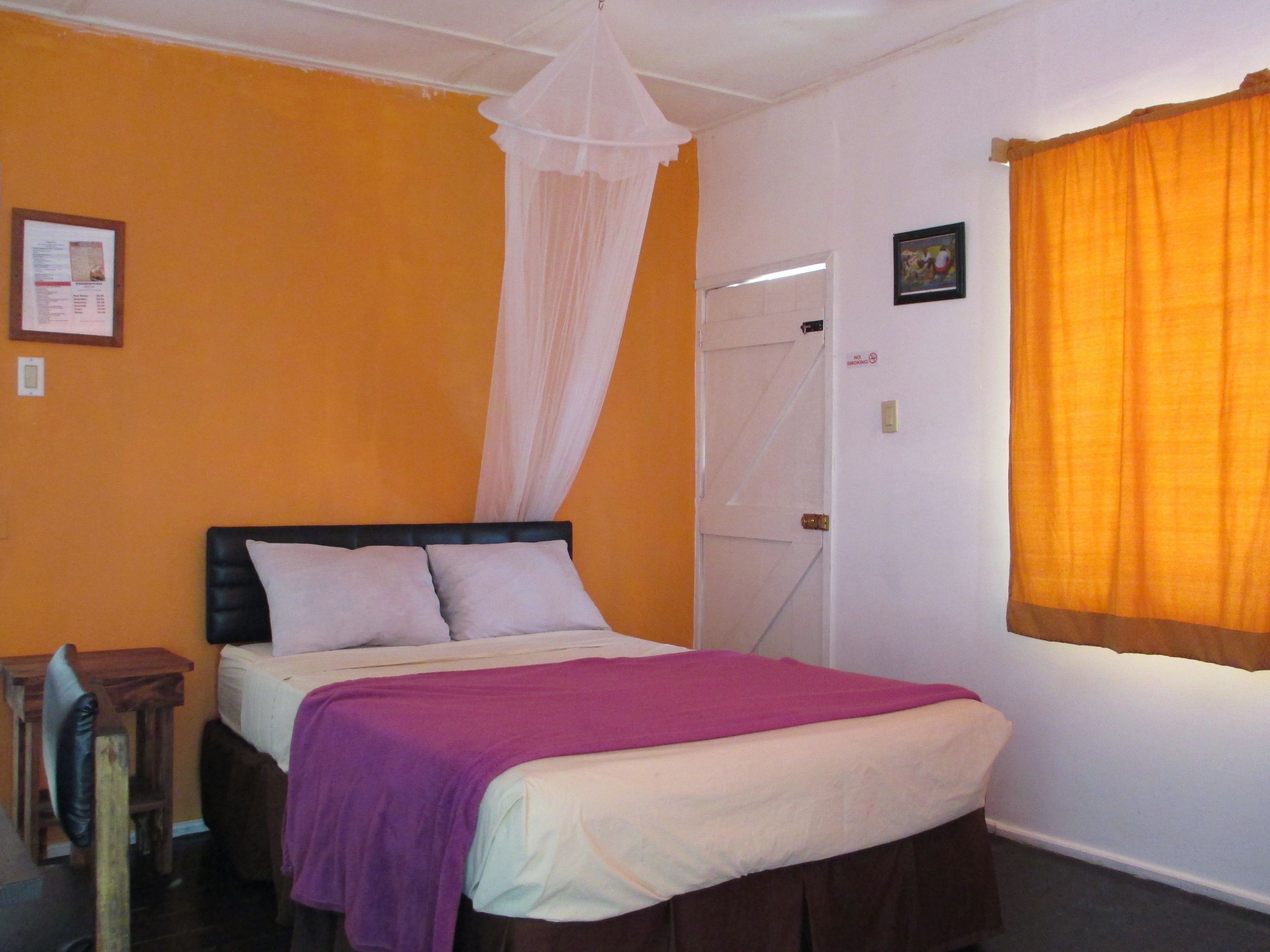 A bedroom with an orange wall, a bed with a white headboard, a purple blanket, and a white canopy.