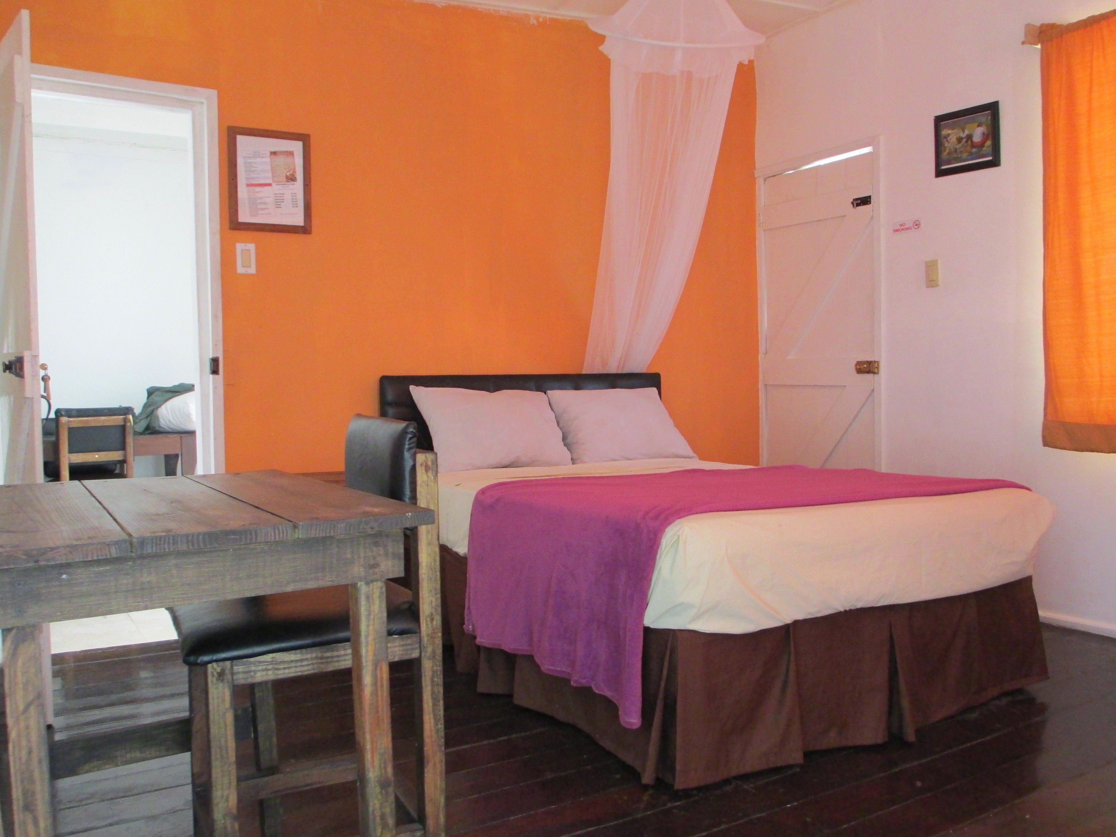 A bedroom with an orange wall, a bed with a purple blanket, a wooden table, and a chair.