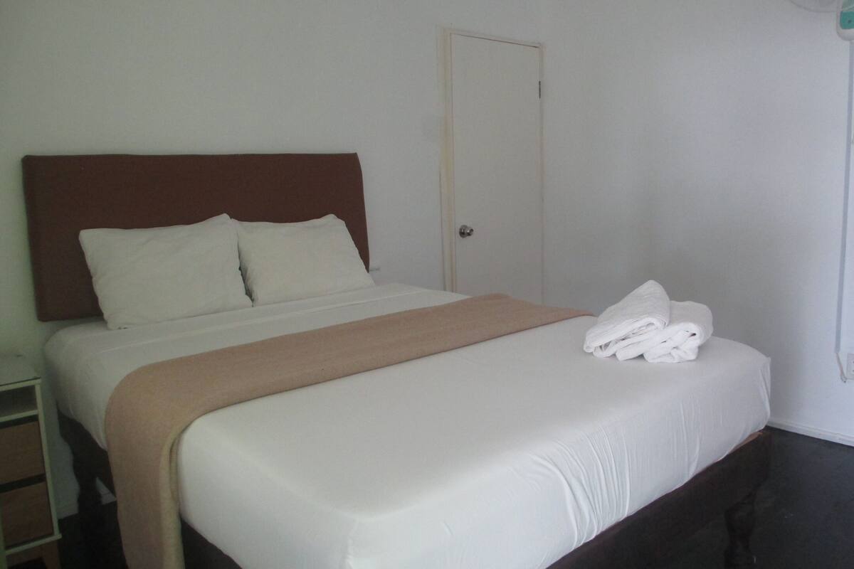 A neatly made bed with white linens and a brown headboard, a closed door, and a stack of white towels on the bed.