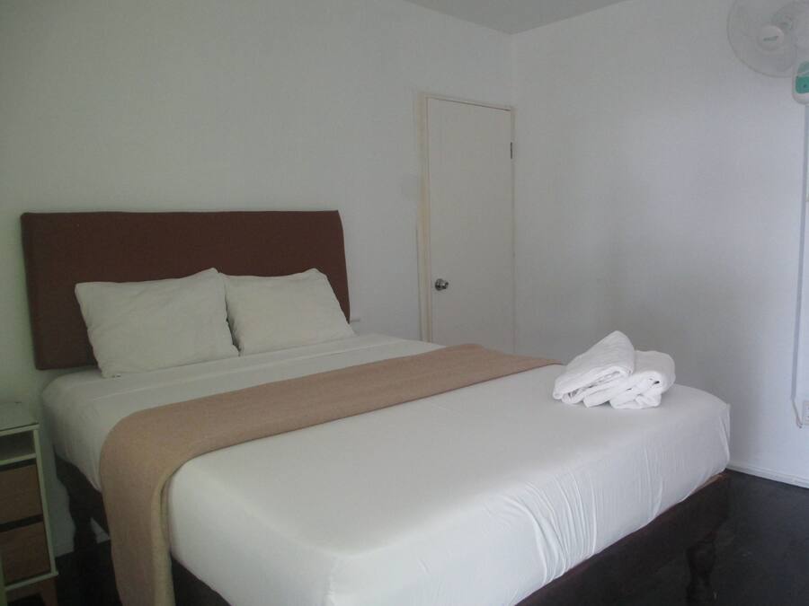 A neatly made bed with white linens and a brown headboard, a closed door, and a stack of white towels on the bed.