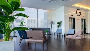 A modern lobby with a high ceiling, large windows, and a ceiling fan. There are seating areas with sofas and chairs, and a large 'G' sign above the elevators.