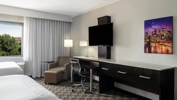 Habitación de hotel con dos camas, un escritorio, una silla, un televisor y una pintura de un paisaje urbano.