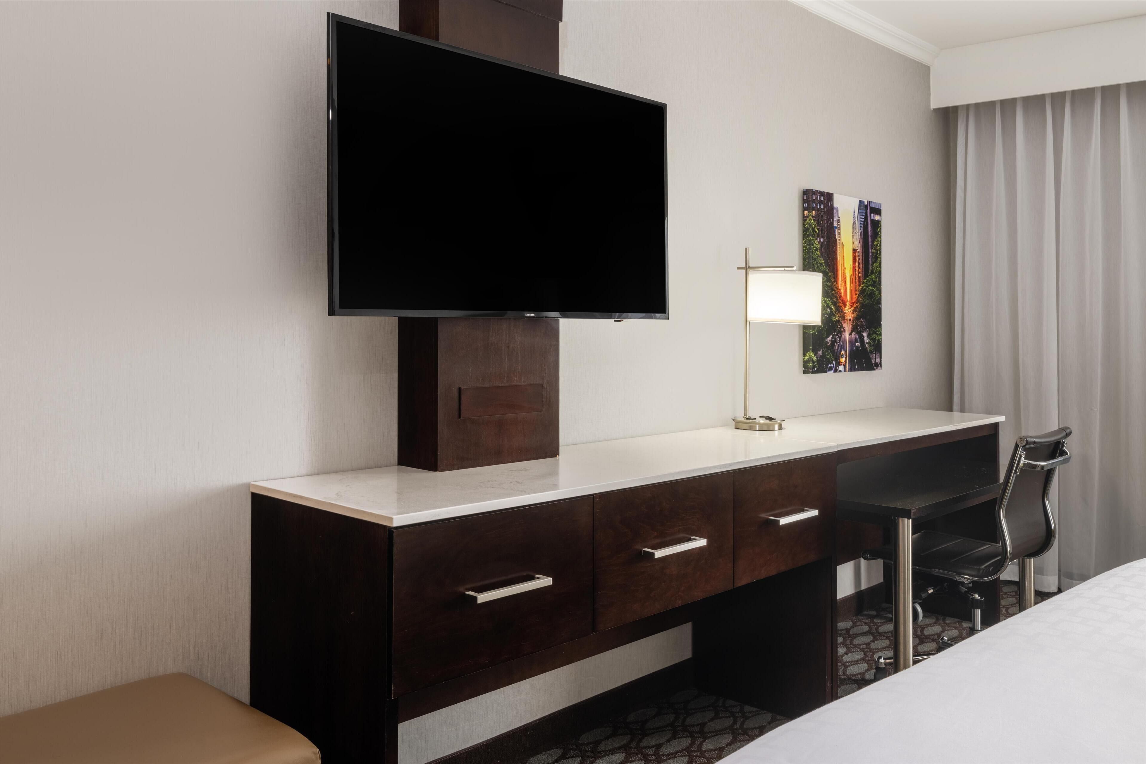 A hotel room with a flat-screen TV mounted on a dark wooden dresser, a desk with a chair, and a bed.