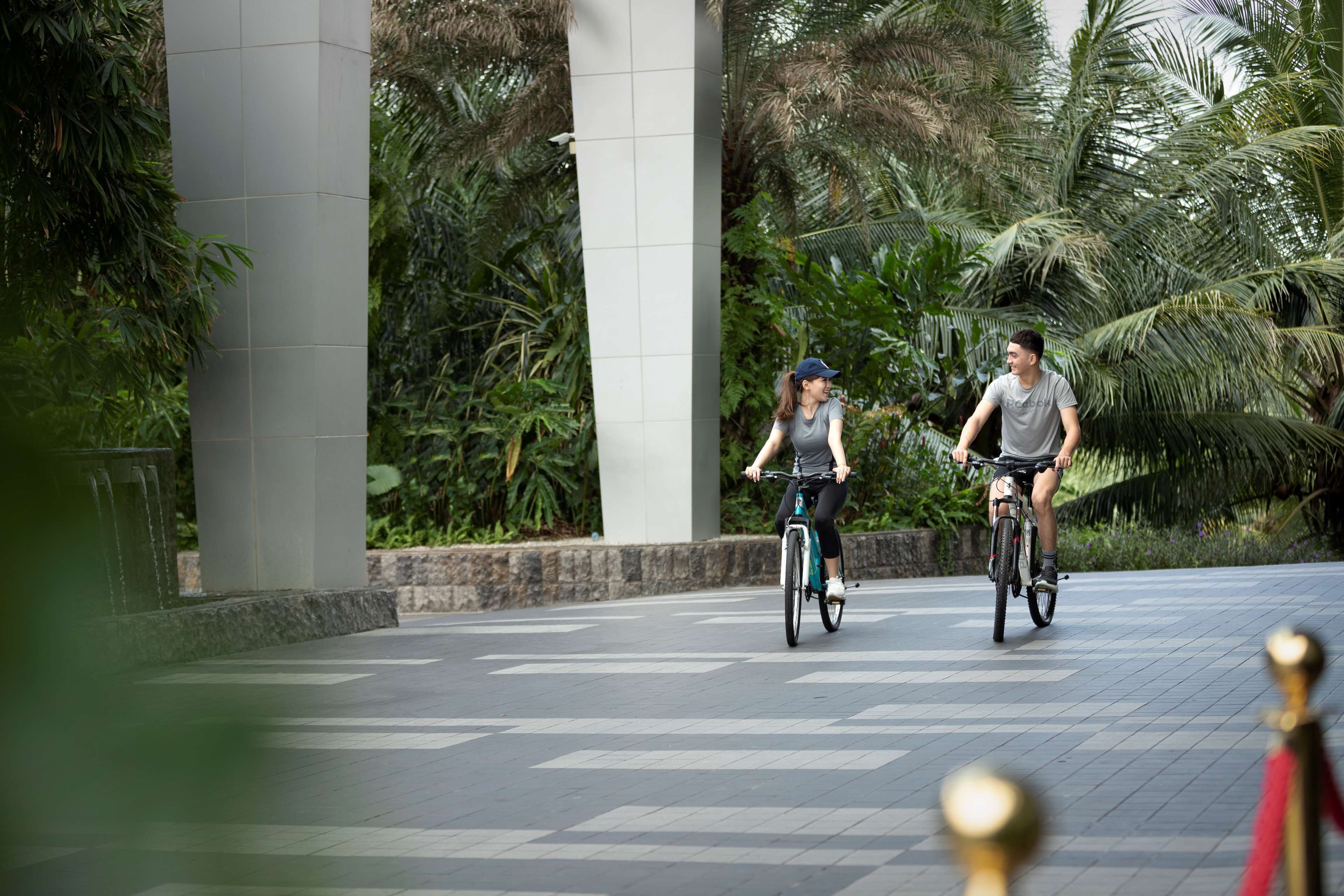 Dos personas montando en bicicleta por un camino pavimentado, rodeado de exuberante vegetación y una gran estructura de columnas.