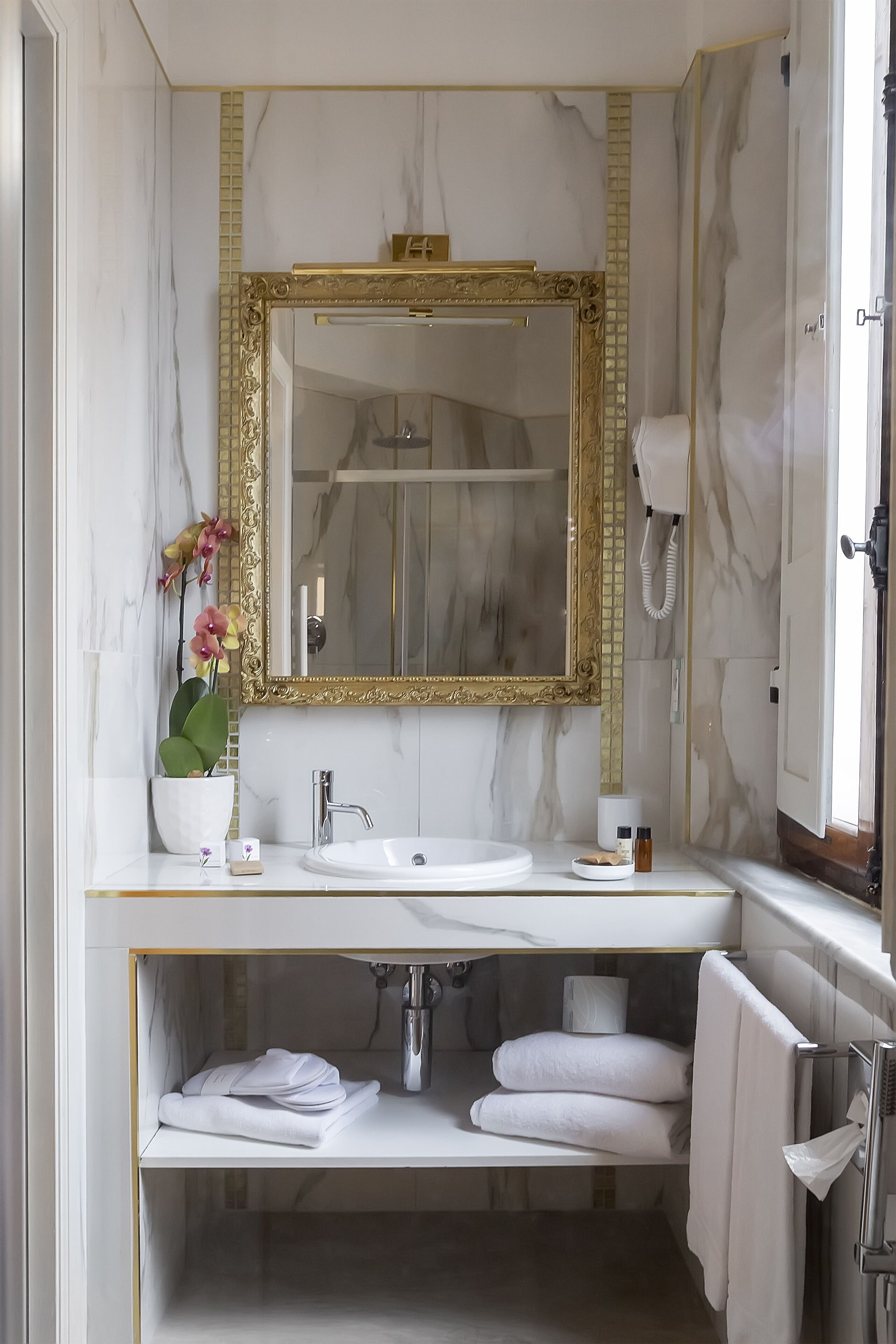 A bathroom with a marble vanity, a gold-framed mirror, a potted orchid, and a towel rack.