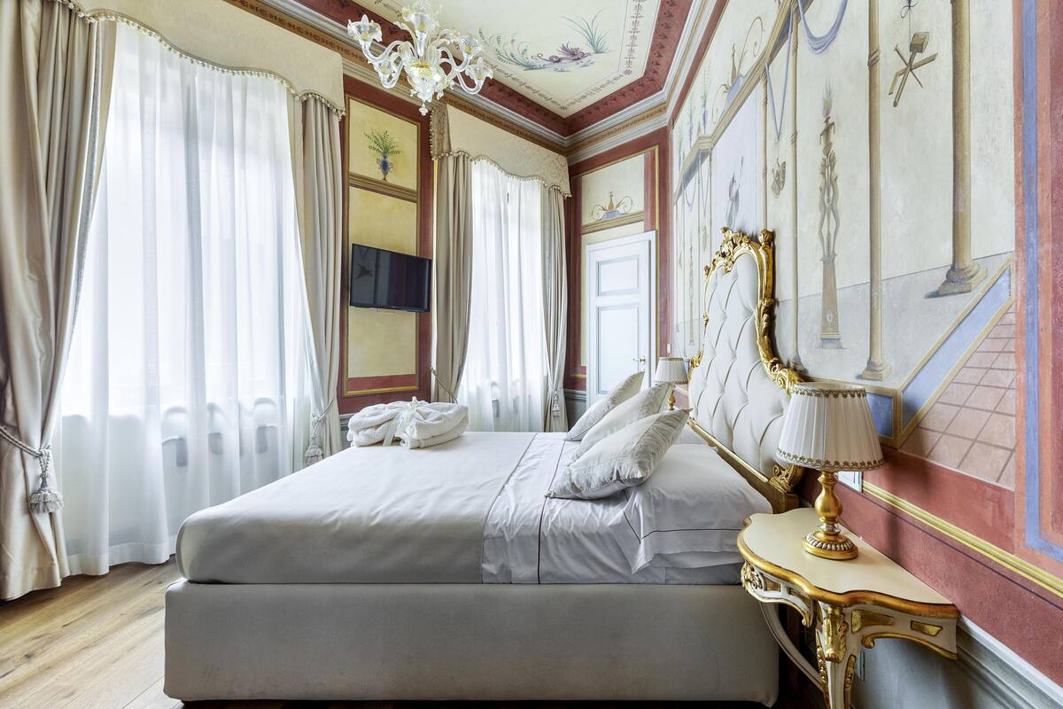 A large bed with white linens, a gold-framed nightstand with a lamp, and a television on the wall.