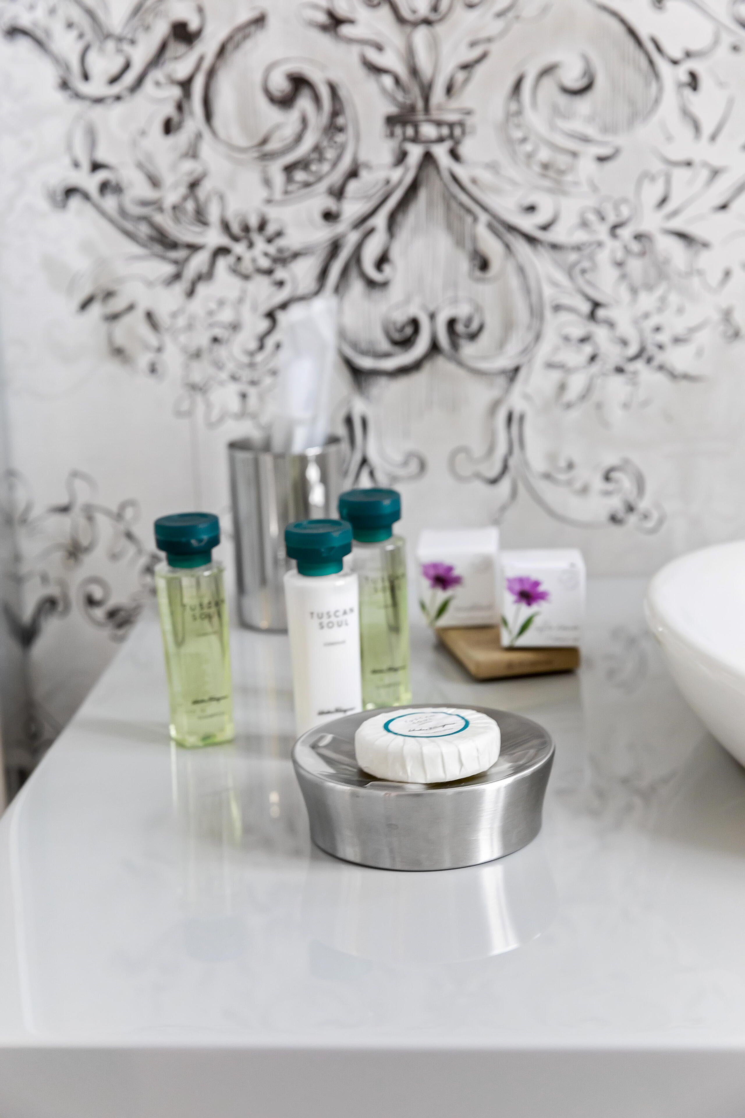 A bathroom vanity with a marble countertop, a metallic soap dish, and toiletries including a bottle of Tuscaran Soul body wash, a green bottle, and a bar of soap.