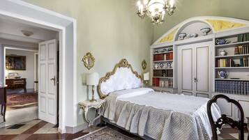 Een slaapkamer met een groot bed, een kroonluchter, een boekenkast en een deur naar een andere kamer.