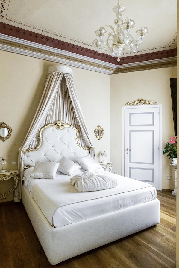 A four-poster bed with a canopy, a chandelier, and a white door in a room with wooden flooring and decorative moldings.