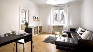 Una sala de estar moderna con un sofá de cuero negro, una mesa de comedor con sillas y vistas a un edificio a través de una ventana.
