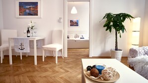 Una habitación con piso de madera, una mesa de comedor con una botella y vasos, una silla, una cama con ropa blanca y una lámpara colgante, además de una planta en maceta.