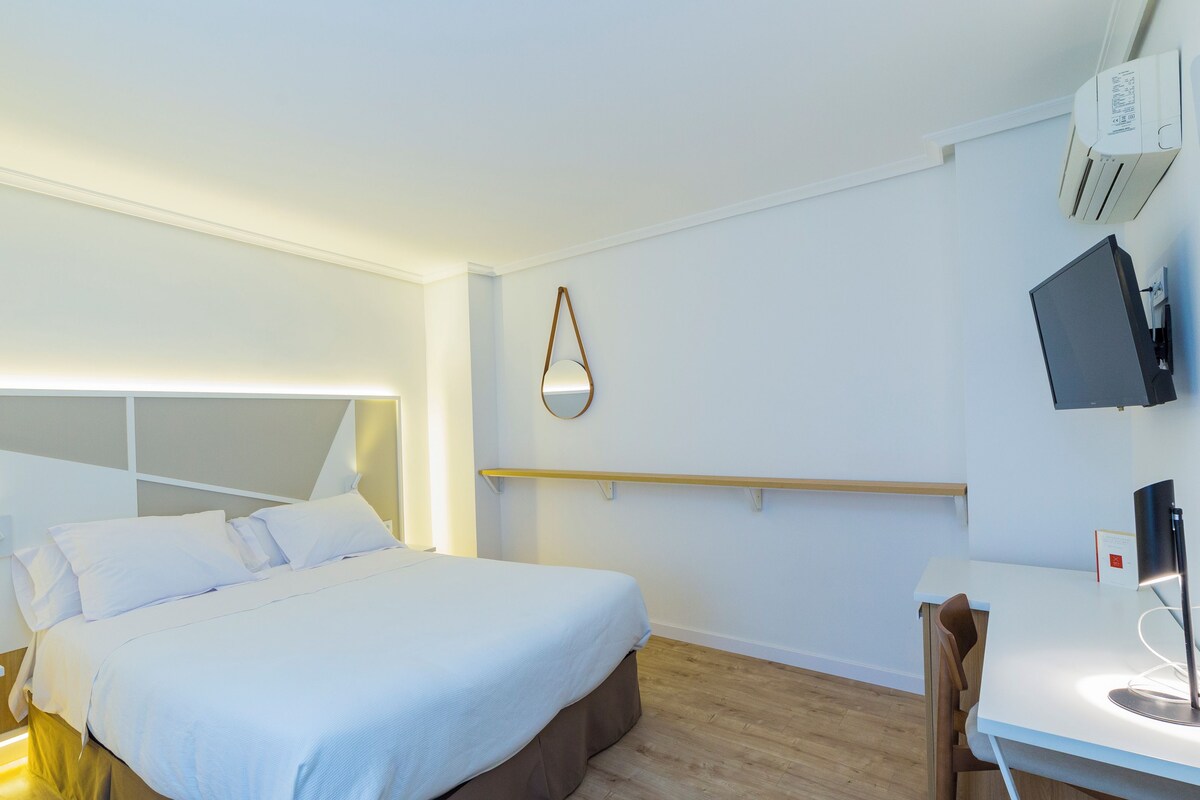 A modern hotel room with a large bed, bedside table, and a desk with a chair.