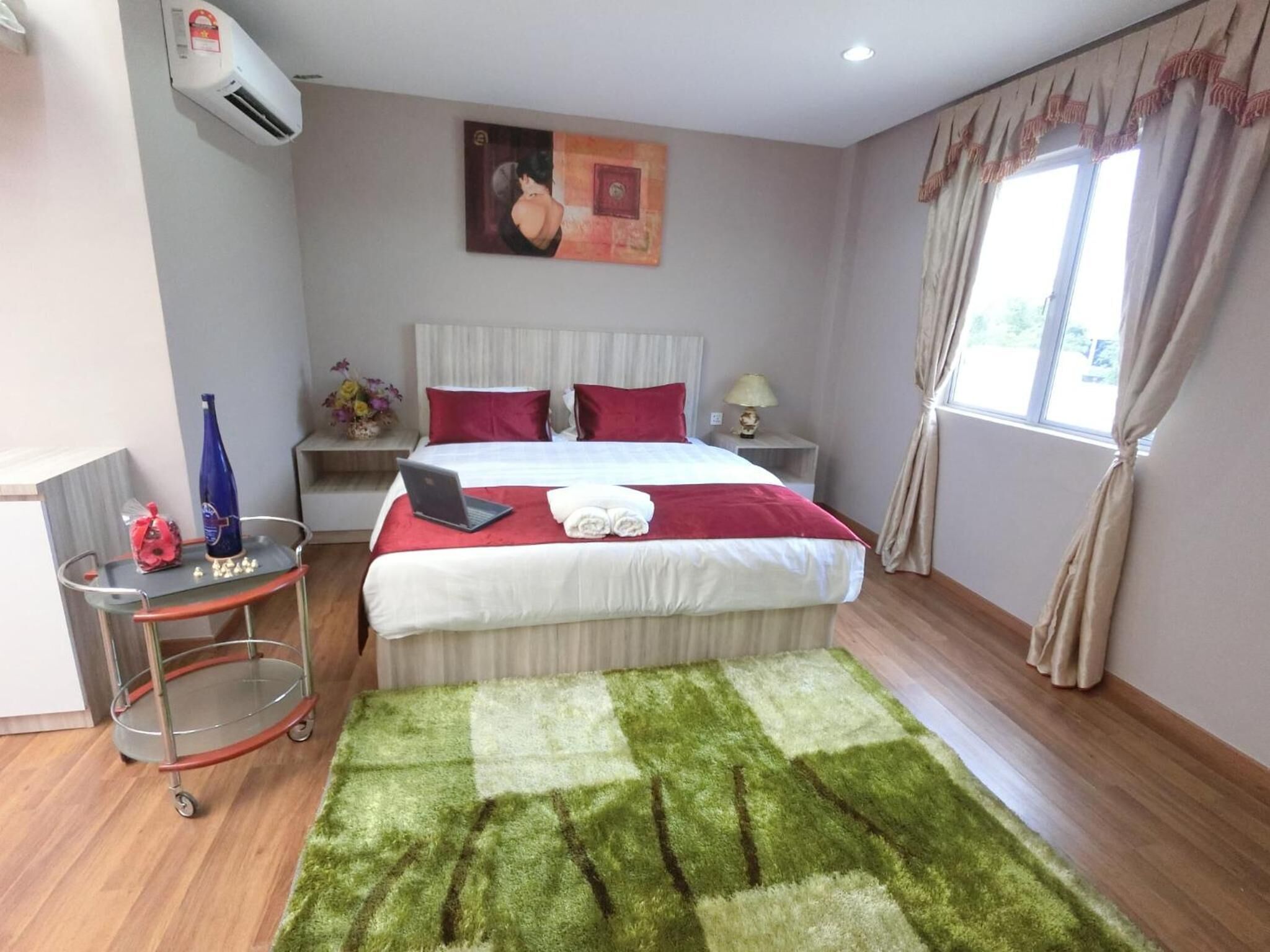 A hotel room with a bed, a desk with a laptop, a small table with a vase and a bottle, and a window with curtains.