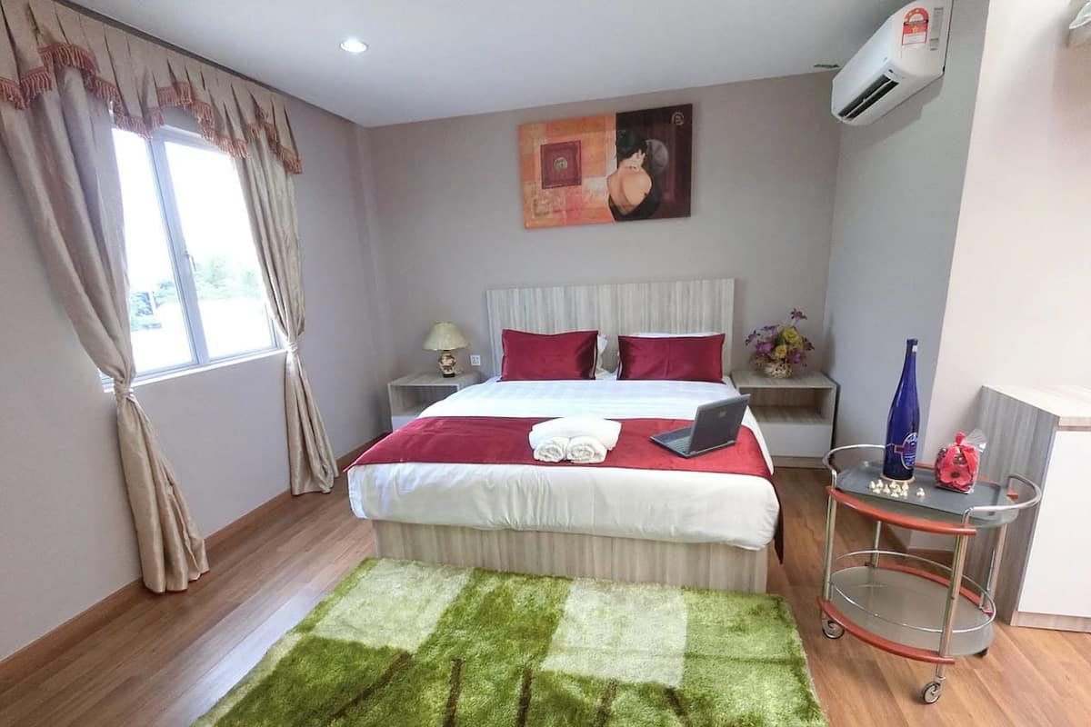 A hotel room with a bed, a green and white rug, a nightstand, a lamp, a painting on the wall, and a cart with a vase.