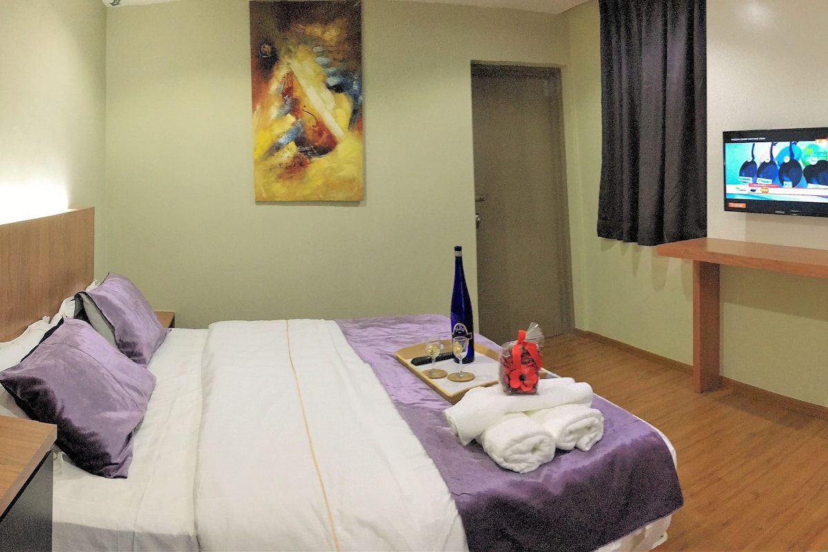 A hotel room with a bed, a painting on the wall, a television, and a small table with a bottle and glasses.