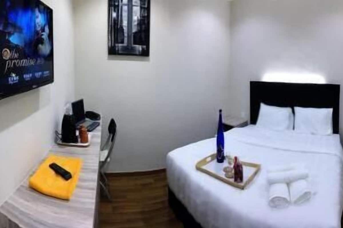 A hotel room with a bed, a bedside table with a remote control, a tray with a bottle and glasses, and a TV mounted on the wall.