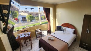 A hotel room with a bed, a small table with a chair, a wardrobe, a window with a view of the street, and a mini-fridge.