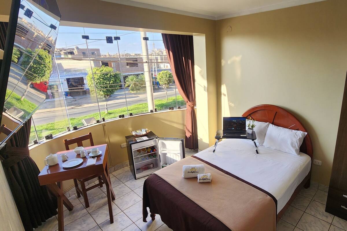 A hotel room with a bed, a small table with a chair, a wardrobe, a window with a view of the street, and a mini-fridge.