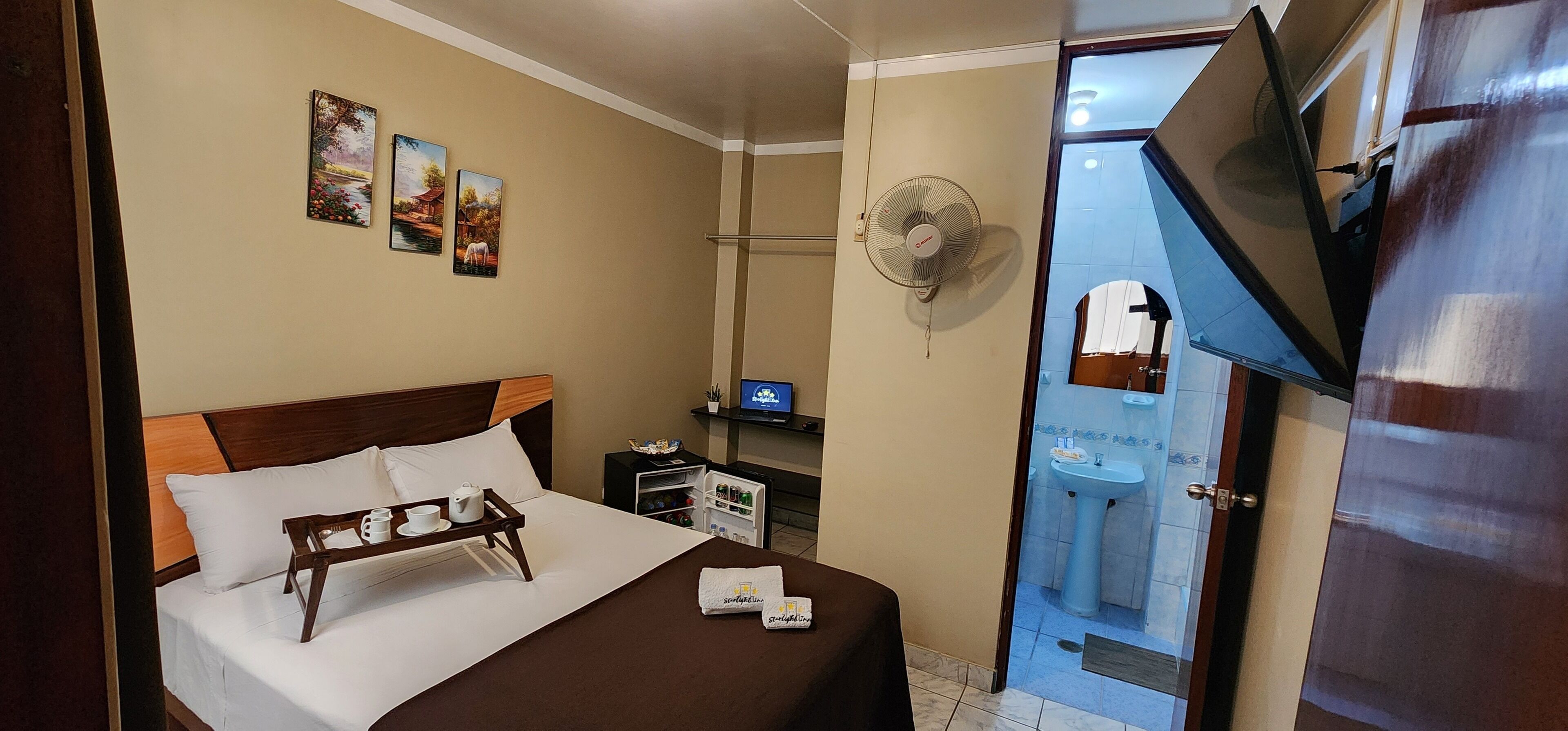 Una habitación de hotel con cama, mesita de noche con bandeja, televisor, ventilador y baño visible a través de una puerta abierta.