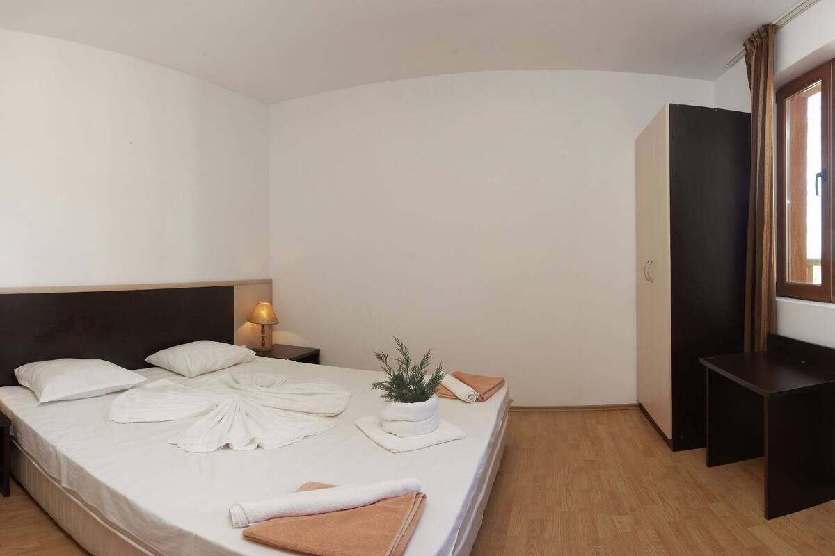 A hotel room with a bed, a nightstand with a lamp, a potted plant, and a wooden floor.