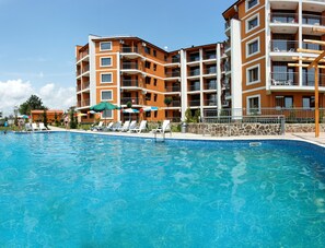 Une piscine entourée de chaises longues et de parasols, elle-même bordée d’un immeuble à plusieurs étages doté de balcons.