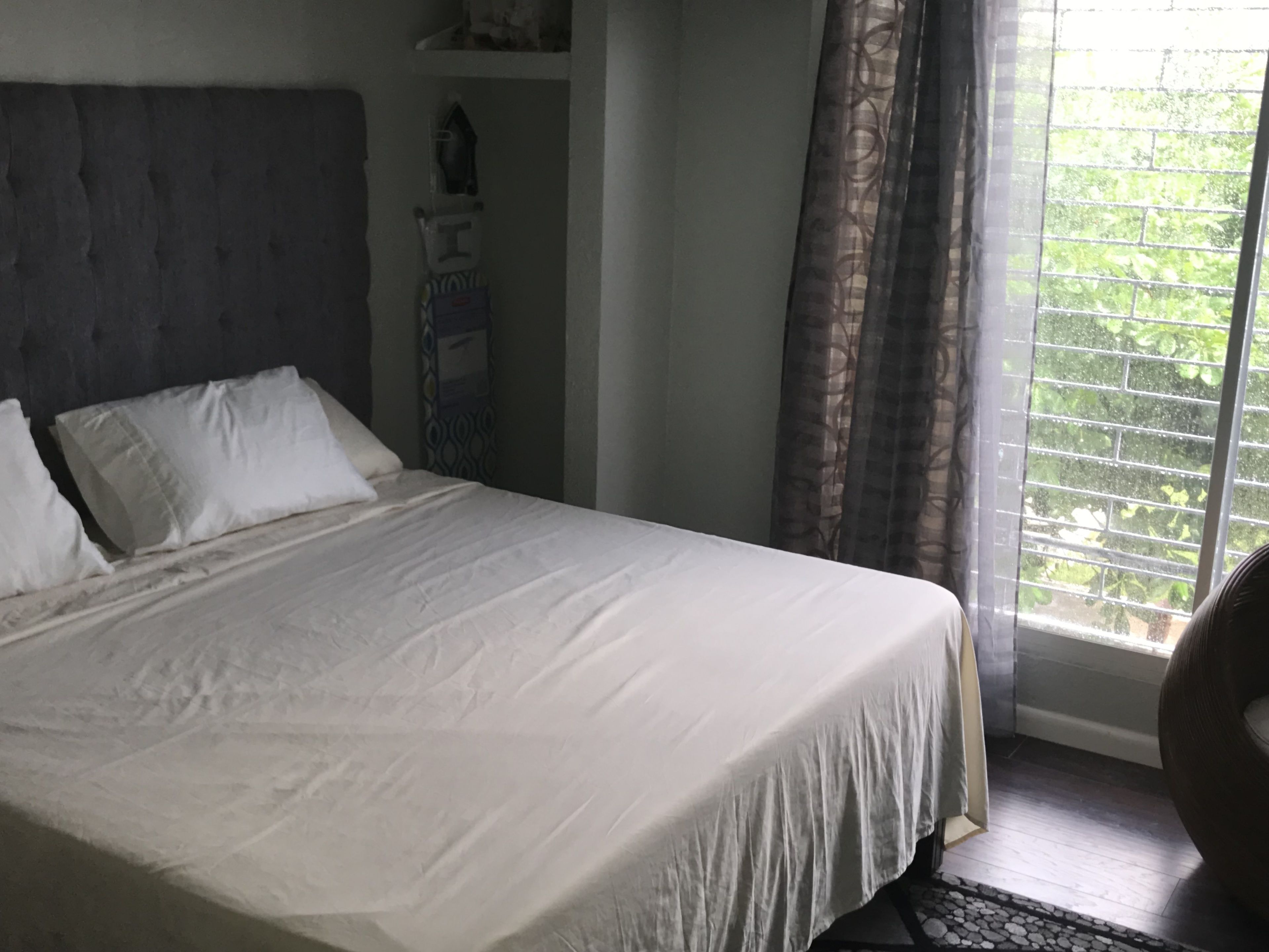 Ein Schlafzimmer mit einem großen Bett, einem Kopfteil, einem Nachttisch mit Lampe und einem Fenster mit Vorhängen.