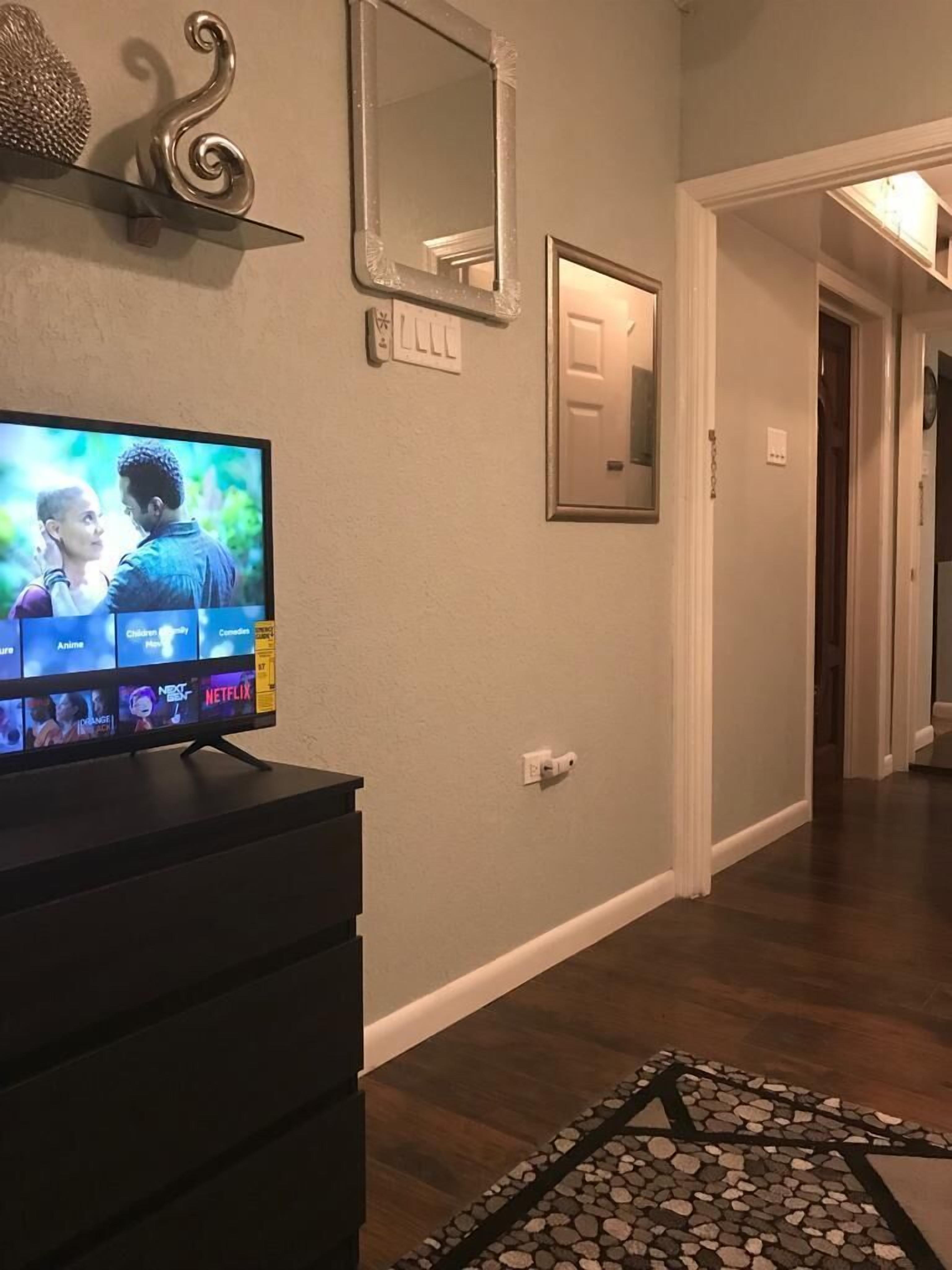 Un salón con un televisor de pantalla plana mostrando la interfaz de un servicio de streaming, un espejo decorativo y una puerta que da a otra habitación.