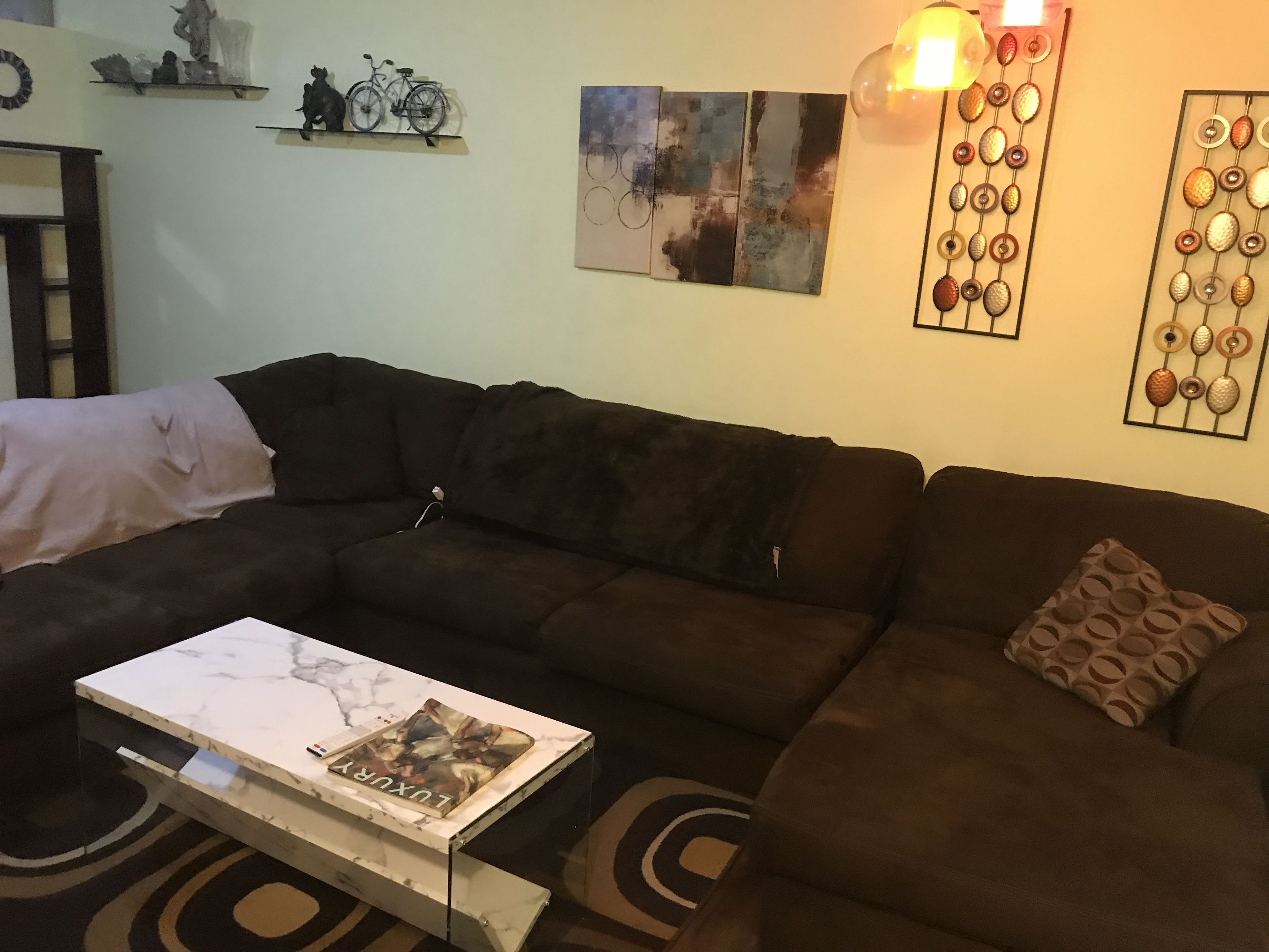 A living room with a sectional sofa, a coffee table with a book, and wall art including a bicycle and abstract paintings.