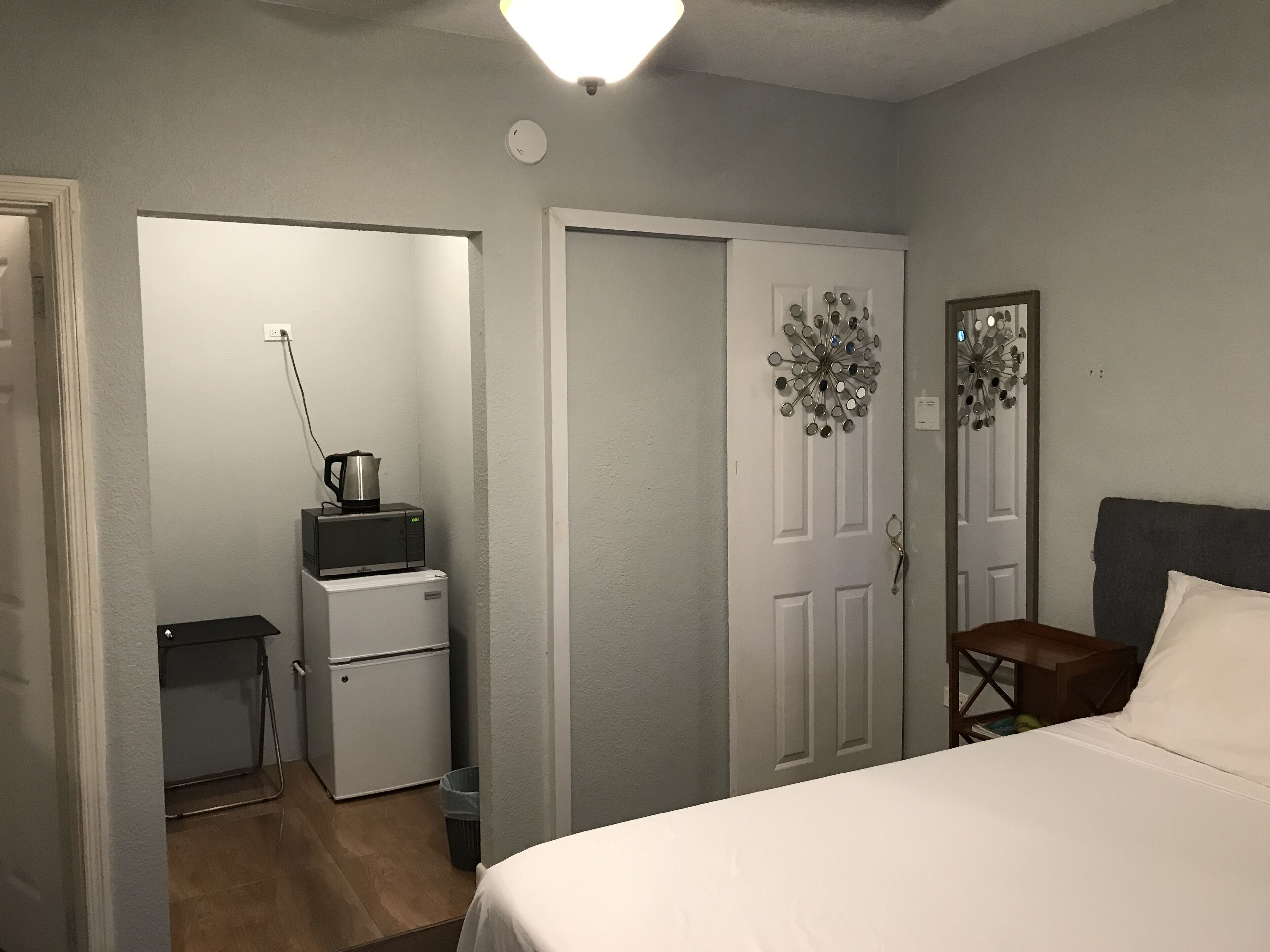 A bedroom with a bed, a desk, a chair, a microwave, a refrigerator, and a door with a decorative mirror.