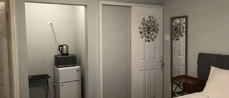 A bedroom with a bed, a desk, a chair, a microwave, a refrigerator, and a door with a decorative mirror.