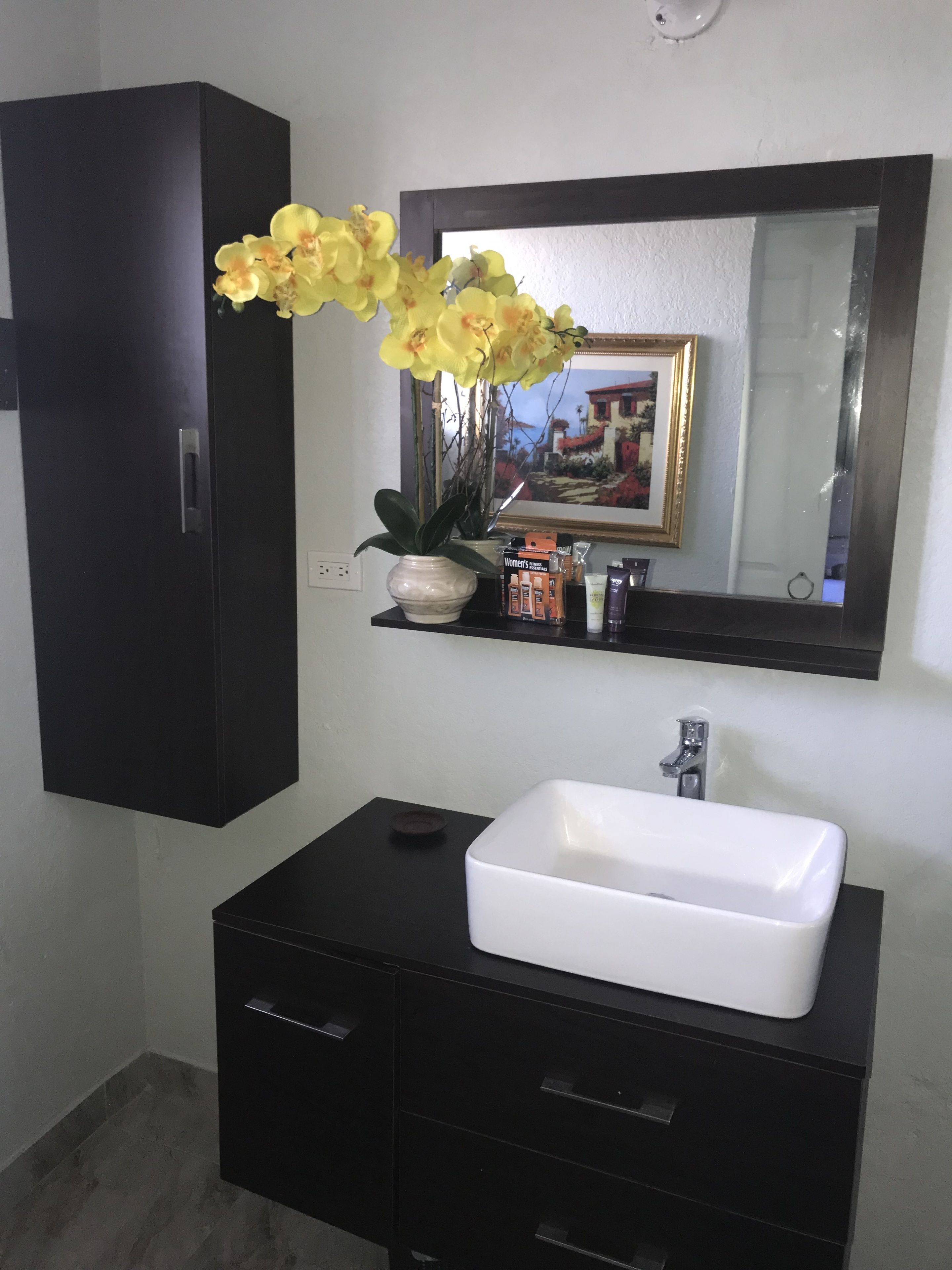 Ein Badezimmer mit einem weißen Waschbecken, dunklen Schränken, einem Spiegel und einer Vase mit gelben Blumen.