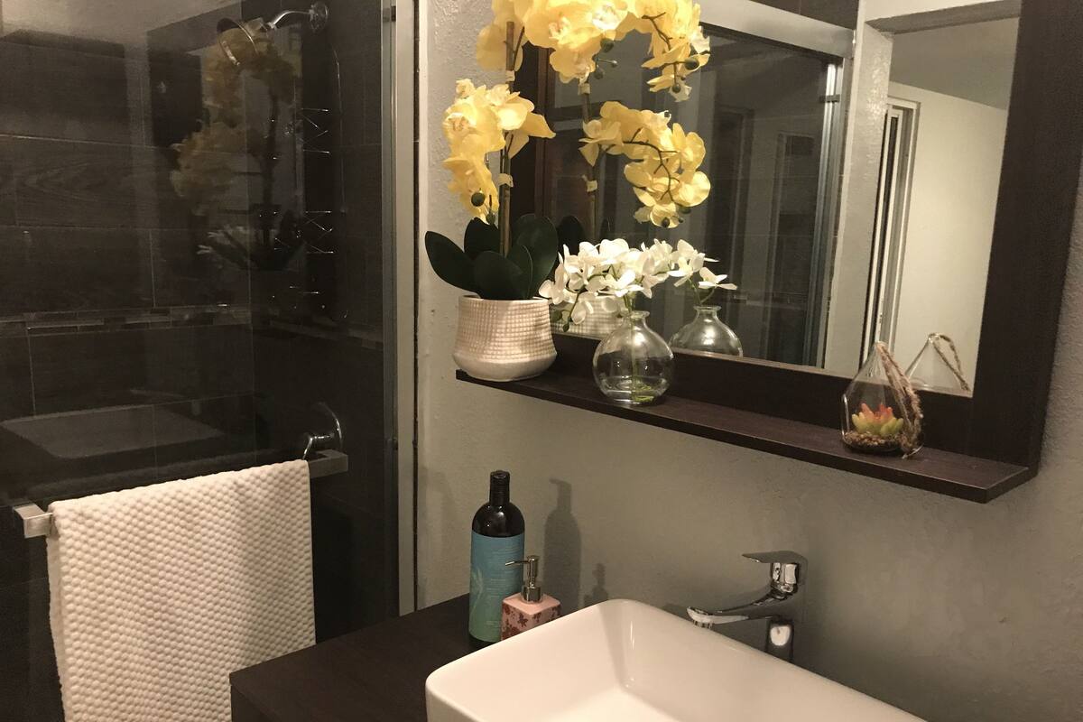 A bathroom with a white sink, a mirror, and a shelf with flowers and a bottle.