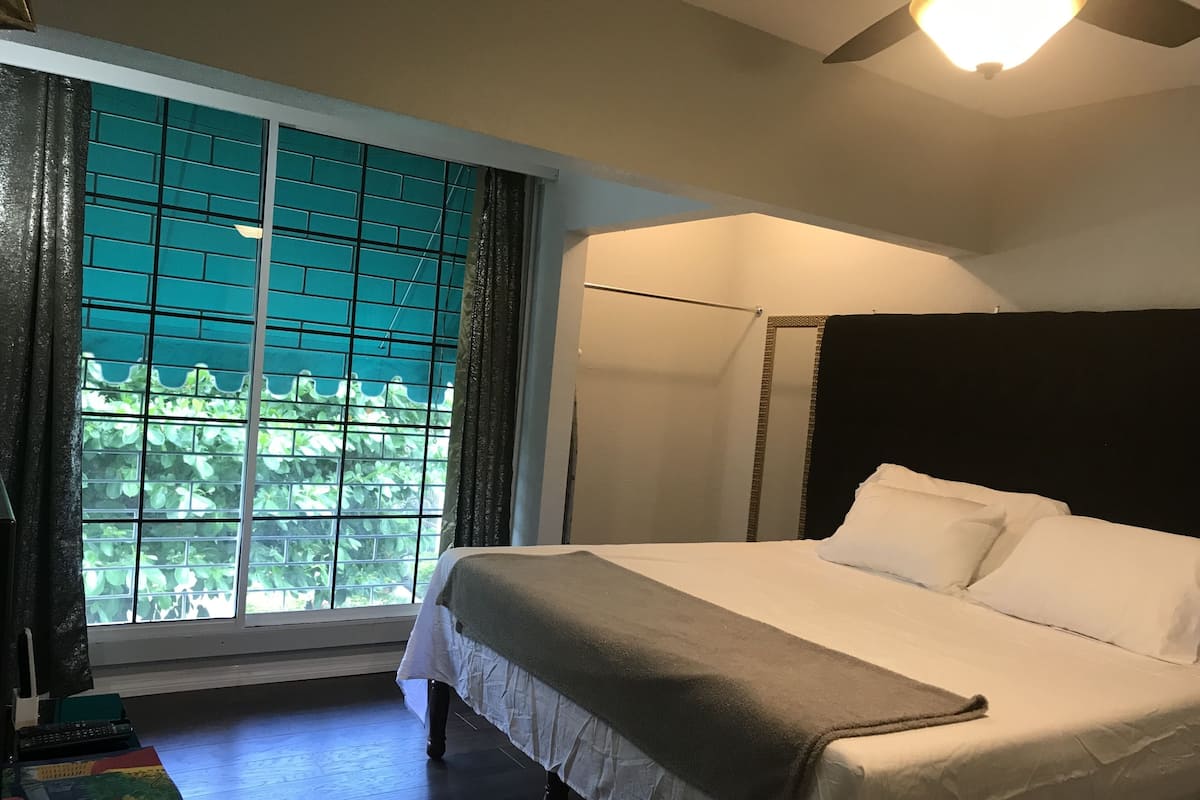 A bedroom with a large window, a bed with white linens, a ceiling fan, and a dark headboard.