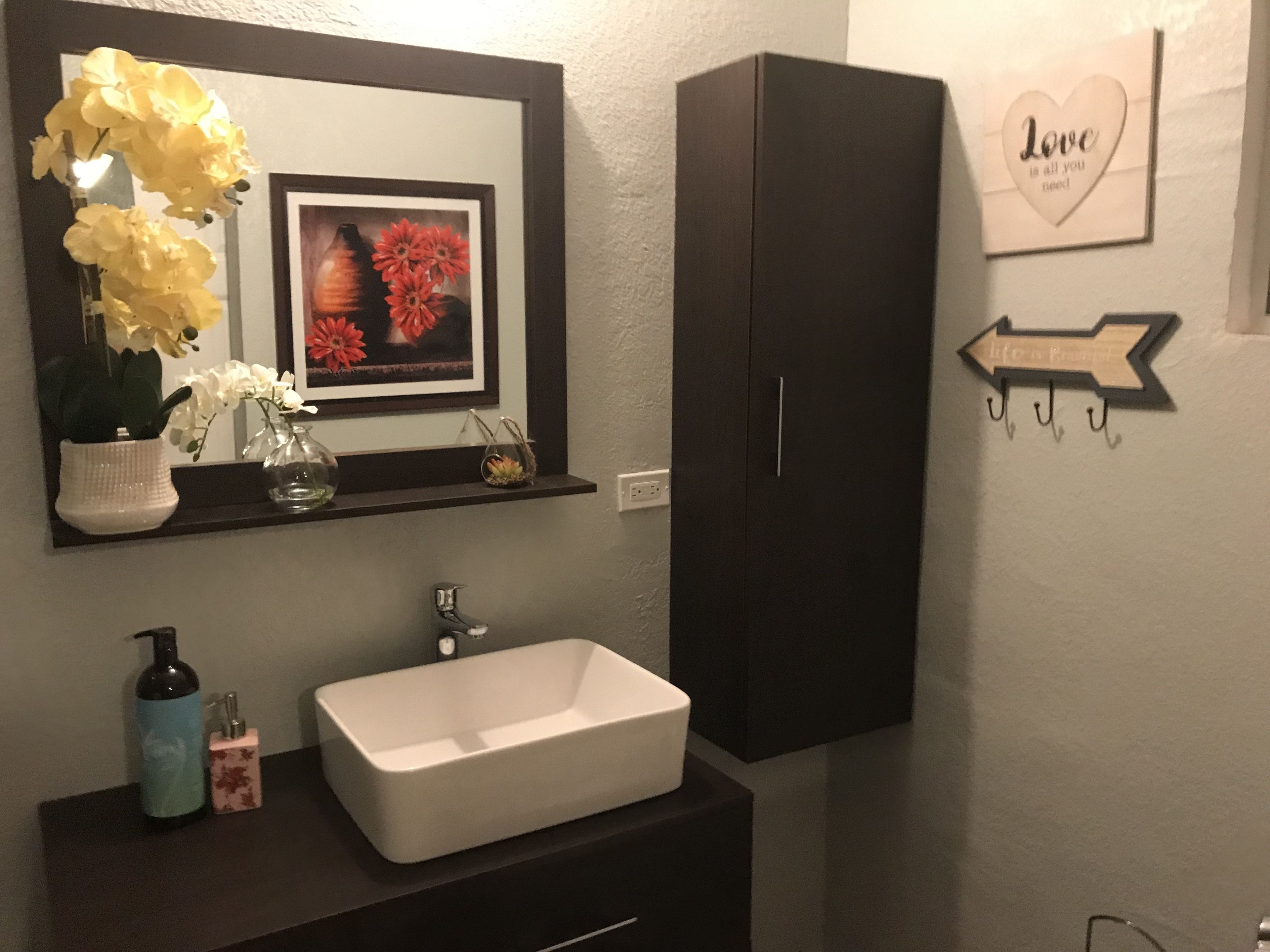 A bathroom with a white sink, dark cabinets, a mirror with a framed picture, and a wall-mounted sign saying "Love is all you need".